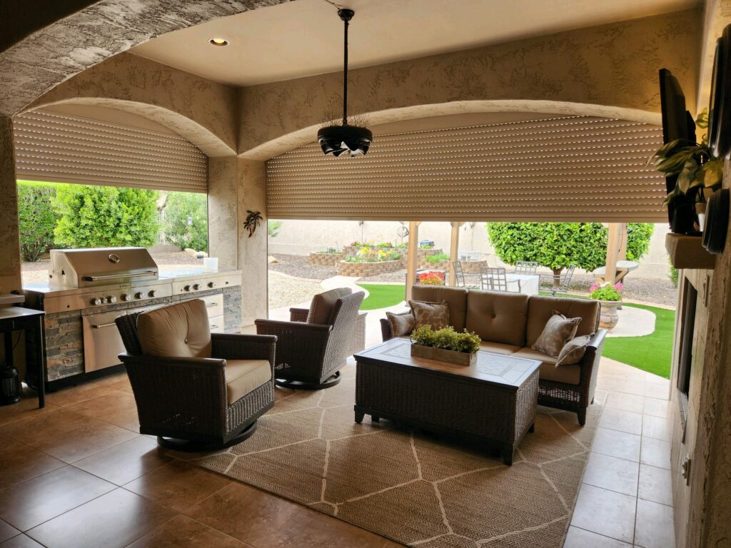 20230413_143329 A covered outdoor living space featuring two wicker chairs and a coffee table with a plant. In the background, a barbecue grill and a seating area are visible, with greenery and landscape design visible through the open windows.
