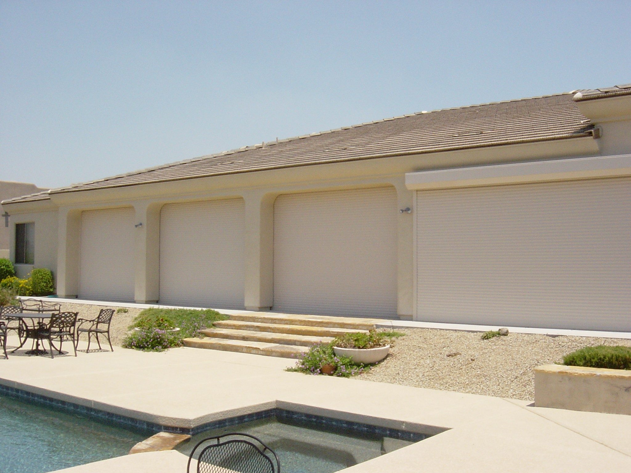 A residential building exterior featuring three closed garage doors with a beige facade. A paved pathway leads to the doors, surrounded by minimal landscaping and a swimming pool in the foreground.