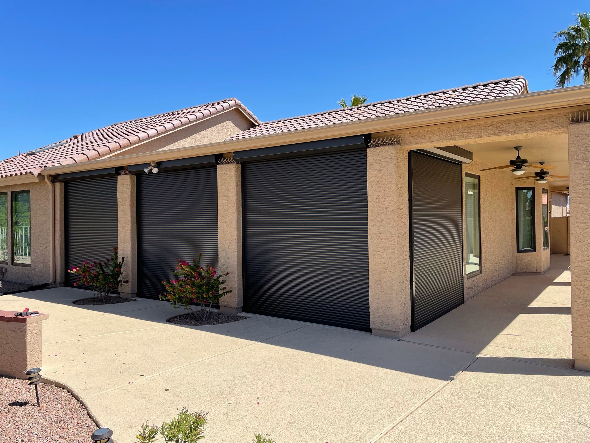 A residential building with three closed roll-up shutters on the exterior, set against a clear blue sky. The ground is paved, and there are small shrubs in front of the building.
