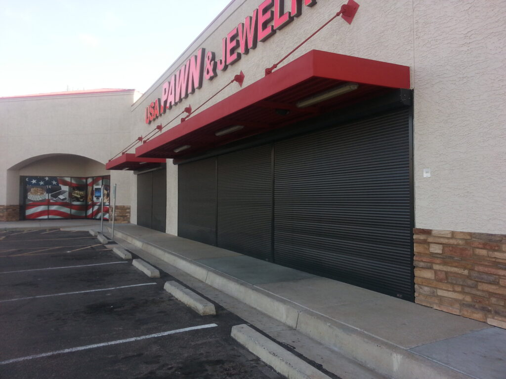 ø; Exterior view of a closed pawn and jewelry shop with rolled-down metal security shutters. The building has a light-colored facade and a red canopy above the entrance. A mural of the American flag is visible on a neighboring wall.