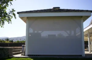 A white patio enclosure with a retractable screen, surrounded by green grass and trees, located in a sunny outdoor area with mountains in the background.