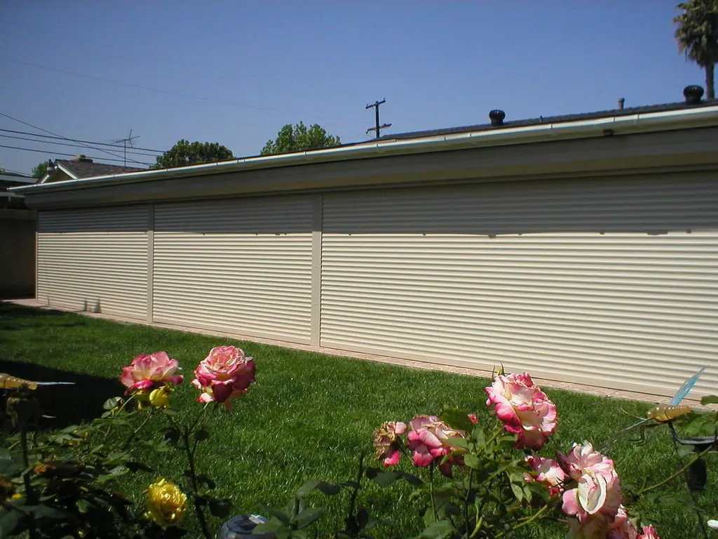 A view of a light-colored garage wall with rolling shutters, surrounded by a green lawn, and pink and yellow roses in the foreground under a clear blue sky.