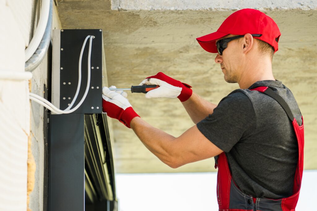 Professional,Residential,Window,Shutters,Instalator,Finishing,Window,Cover,Assembly.,Automatic A worker in a red cap and gloves using a screwdriver on a black electrical panel mounted on a wall. The worker is wearing a black shirt and red overalls, with various cables visible in the background.