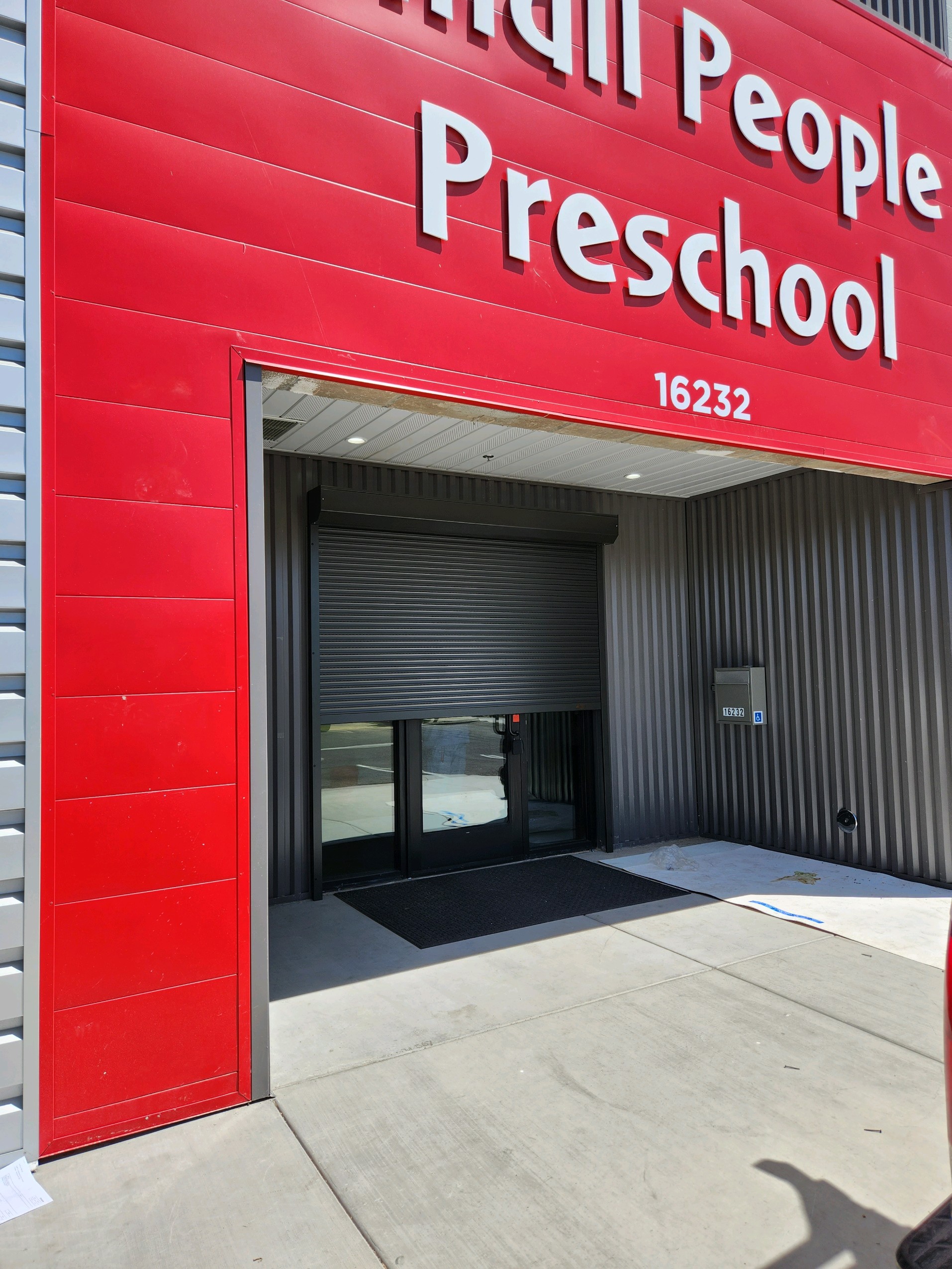 Entrance of Small People Preschool, featuring a red facade and a roll-up gate partially closed. The location number, 16232, is displayed above.