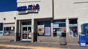The exterior of Mesa Star Food Mart, featuring a prominent sign with the store name. Large windows showcase various advertisements, and a sign indicates available services. A blue newspaper stand is visible on the right, with a trash can in front.