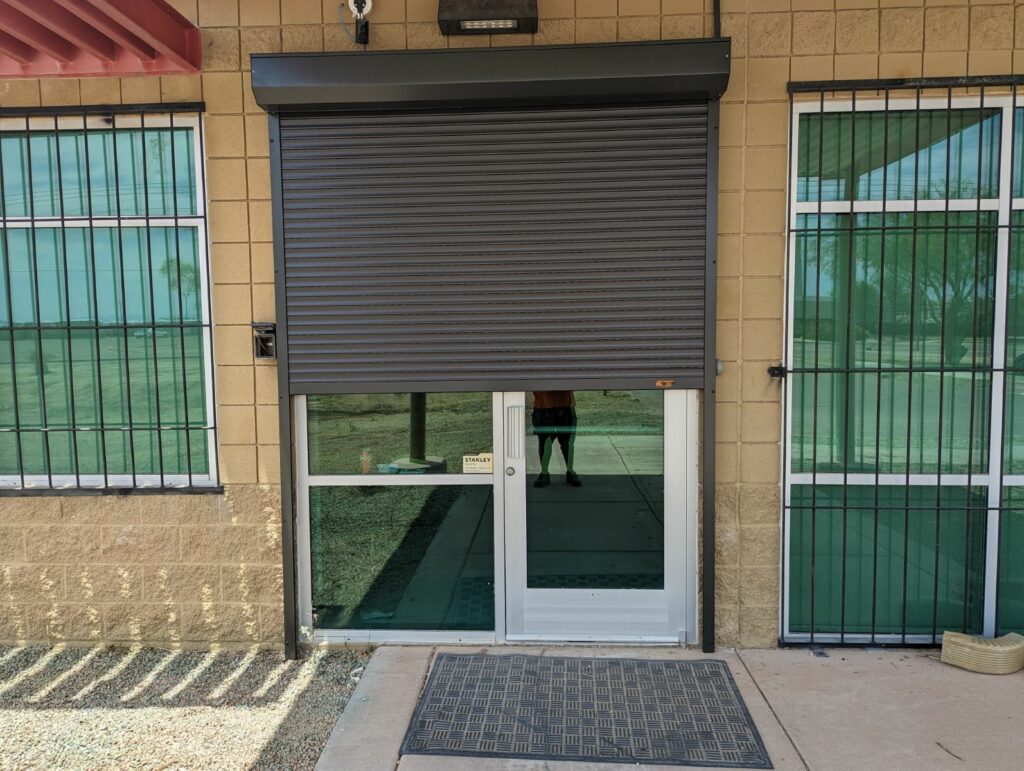 socal-commercial-4 A building entrance featuring a rolling security shutter partially closed over a glass door. The door is framed by metal grilles on either side, with a textured entrance mat in front.