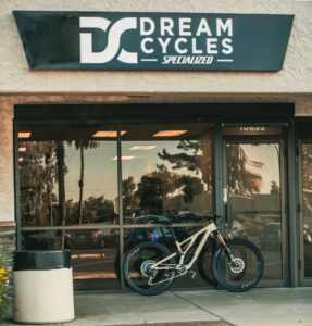 A mountain bike parked outside Dream Cycles, a bike shop featuring a sign with the name and logo above the entrance. Reflections of trees are visible in the store windows.