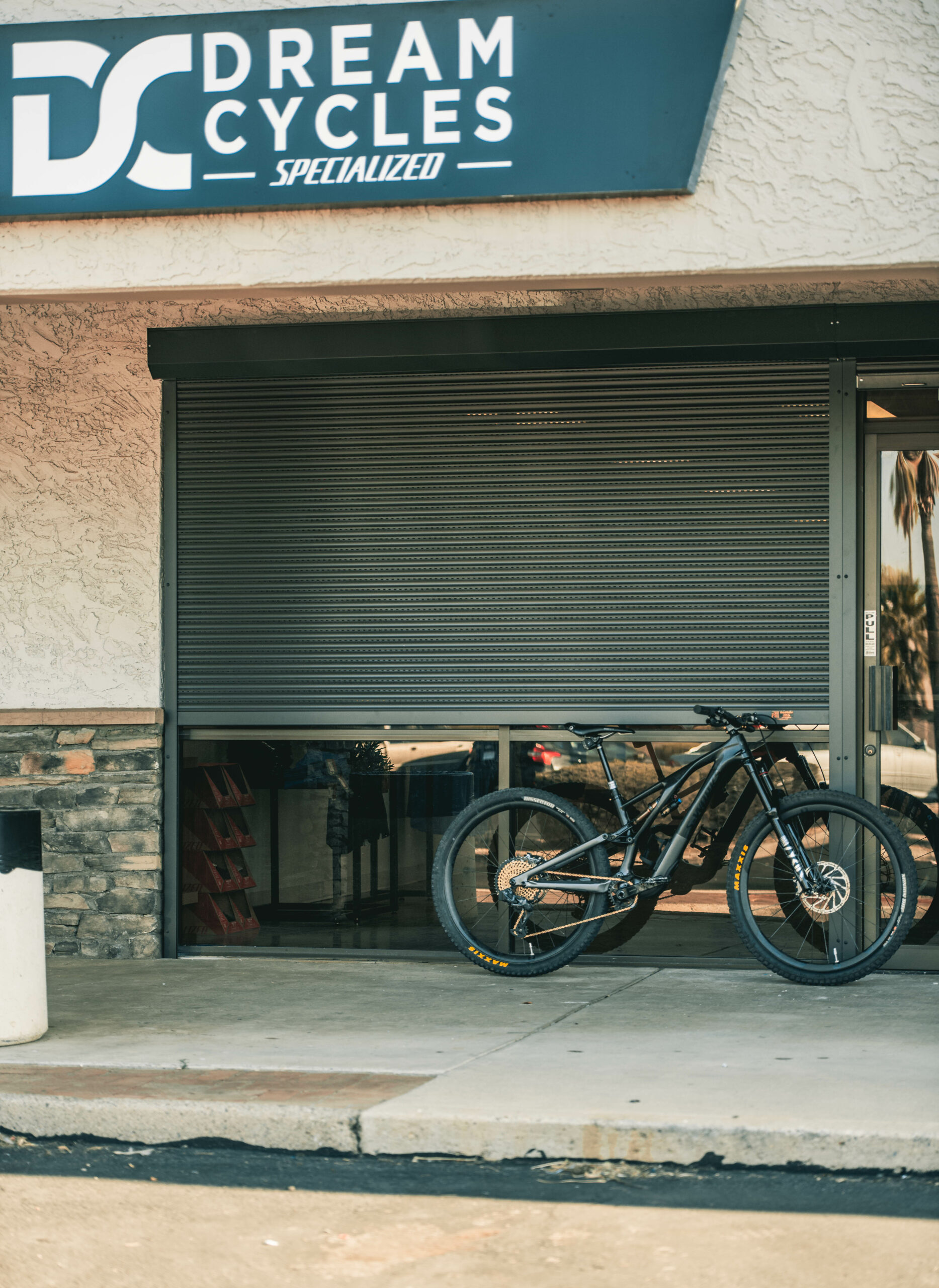 A black mountain bike is parked in front of a shop with a sign that reads 'Dream Cycles Specialized.' The store has a closed shutter and a stone wall design.