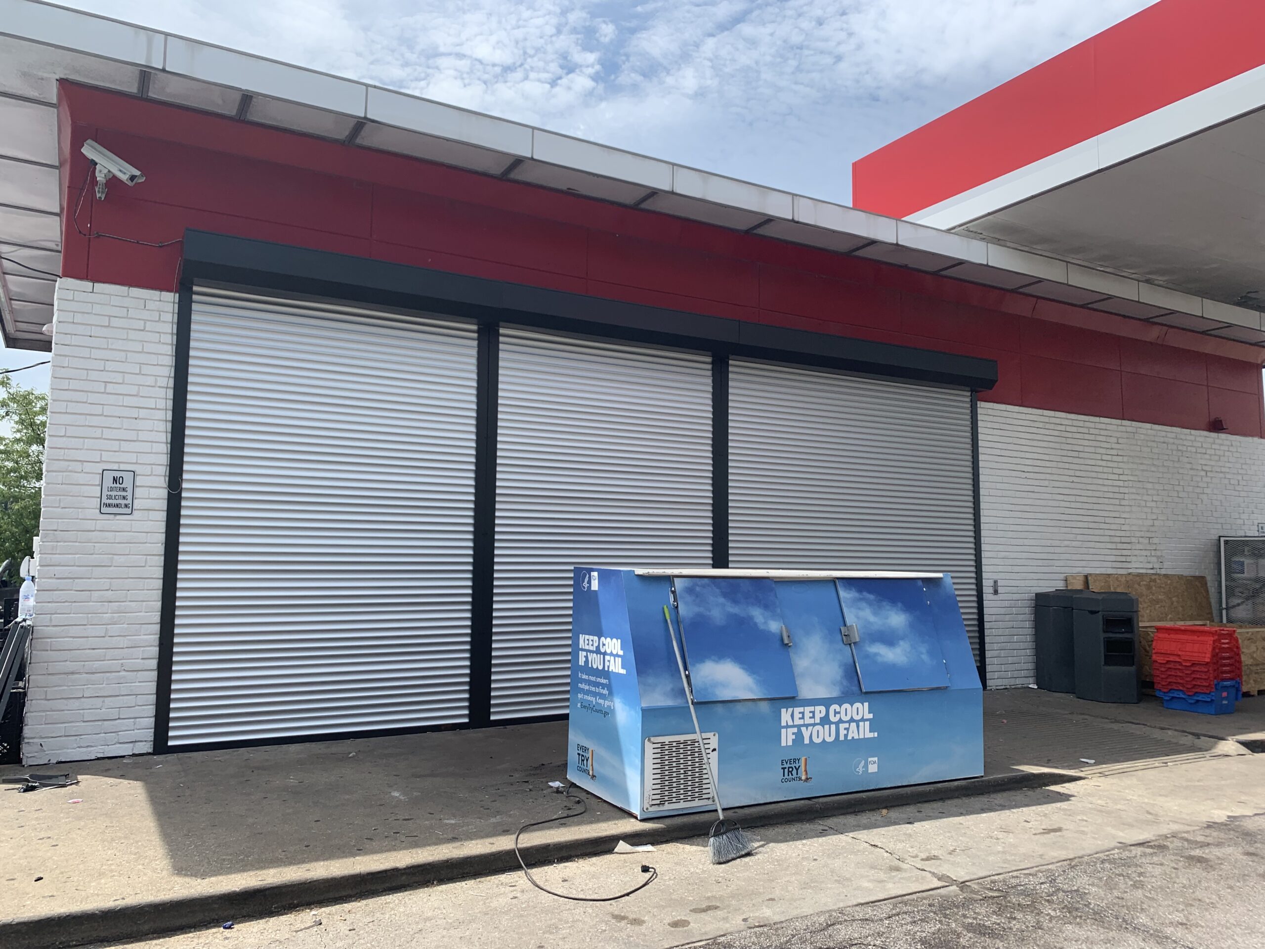 A commercial building with a red and white facade featuring closed metal roll-up doors. In the foreground, a blue utility box is displayed with the text "KEEP COOL IF YOU FAIL." The area is well-lit with a clear sky overhead.