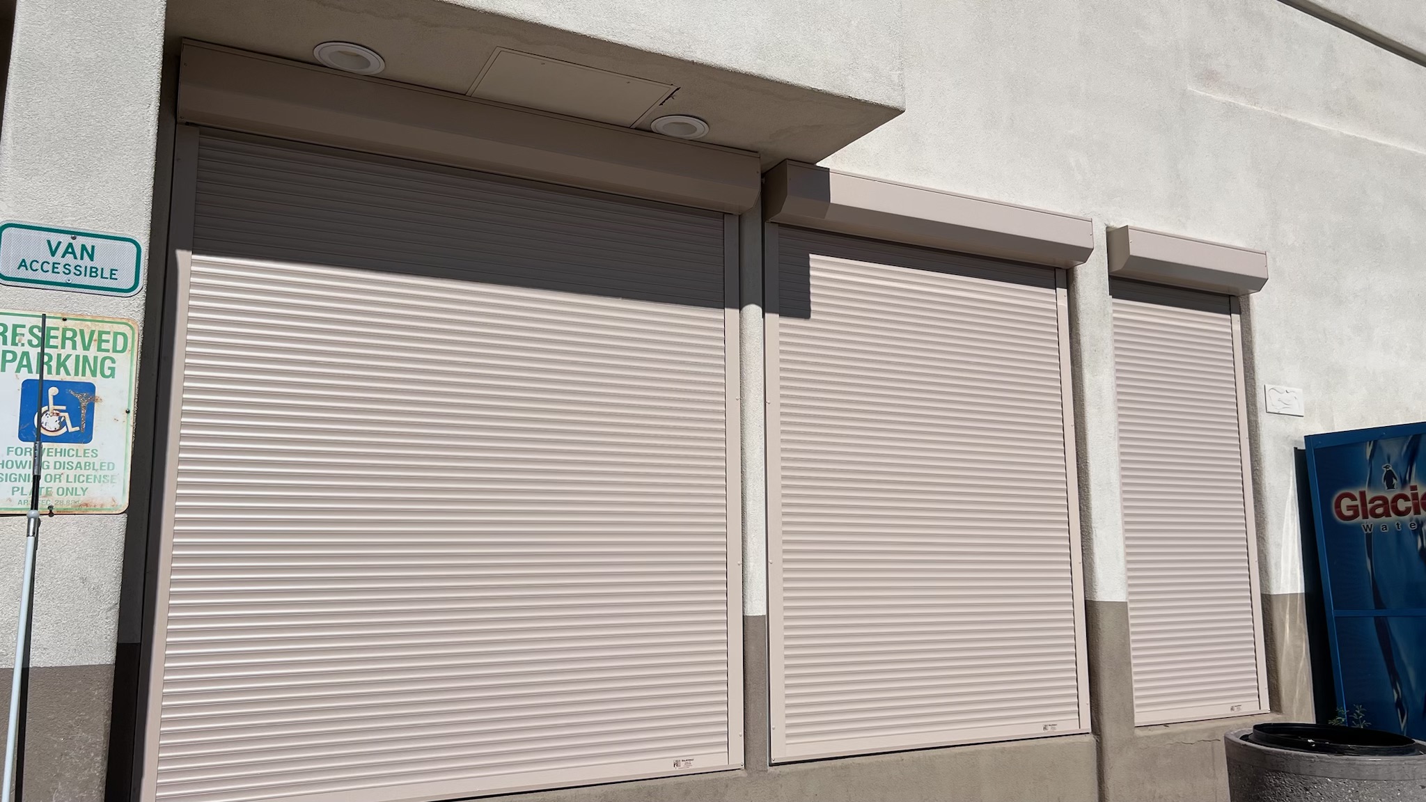 Three closed roll-up shutters on a light-colored wall, with a reserved parking sign for accessible van parking visible on the left.