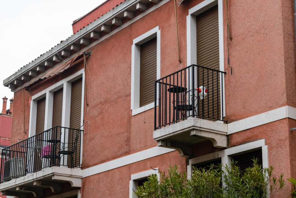Embark,On,A,Captivating,Journey,Through,The,Labyrinthine,Alleyways,Of A pink-stucco building with closed window shutters and two balconies. One balcony has a small table and two chairs, while the other displays a pink item. Greenery is visible below.