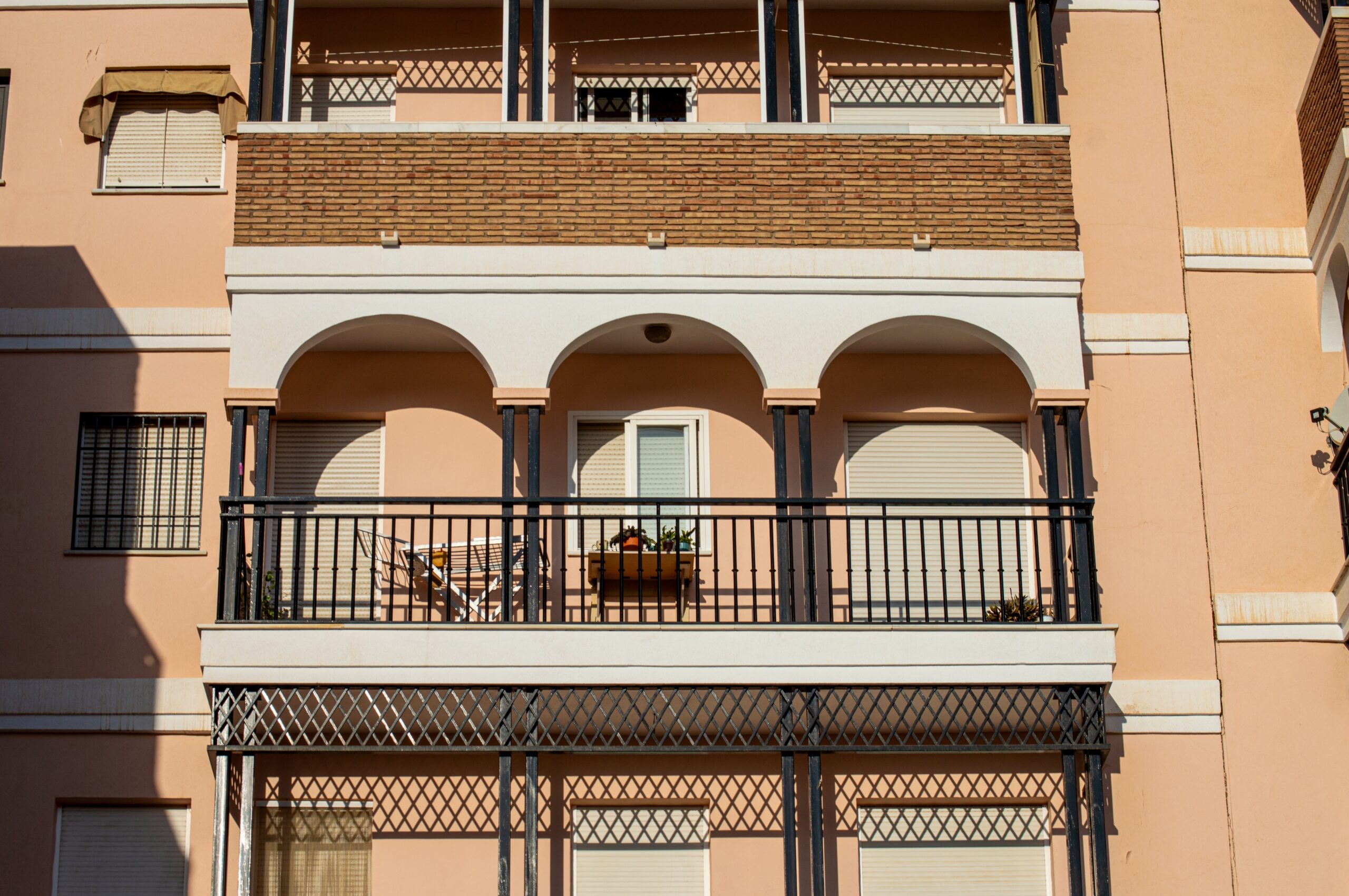 A peach-colored building featuring two balconies with arched railings. One balcony has a potted plant and a chair visible. The windows above are covered with blinds, and the overall architecture has a modern style.