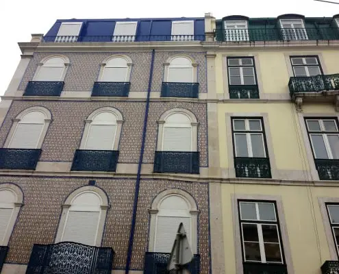 A building facade featuring a combination of blue and beige tiles, with several windows and ornate balconies. The ground level has a white umbrella visible in front, and the structure shows a mix of architectural styles.