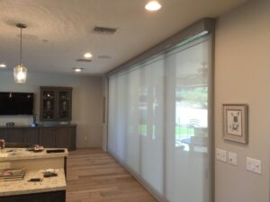 An interior view of a modern kitchen and living room with wooden flooring. A sheer roller shade covers a large window, allowing some light to filter in. The room features a TV, cabinetry, and wall decor.