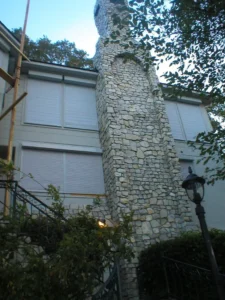 A tall stone chimney attached to a building, surrounded by trees. The building has multiple windows with closed shutters. A black lamp post is visible in the foreground, alongside a winding pathway.