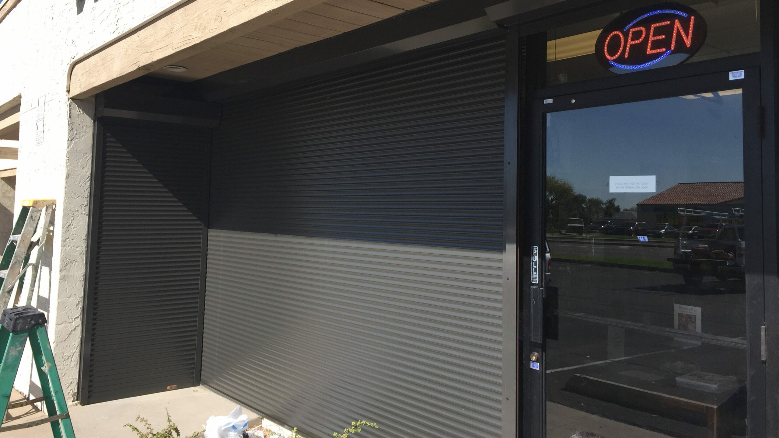 A storefront featuring a gray metal shutter partially closed, with a bright 'OPEN' sign displayed. The entrance area includes a small table with potted plants and part of a ladder visible on the left.