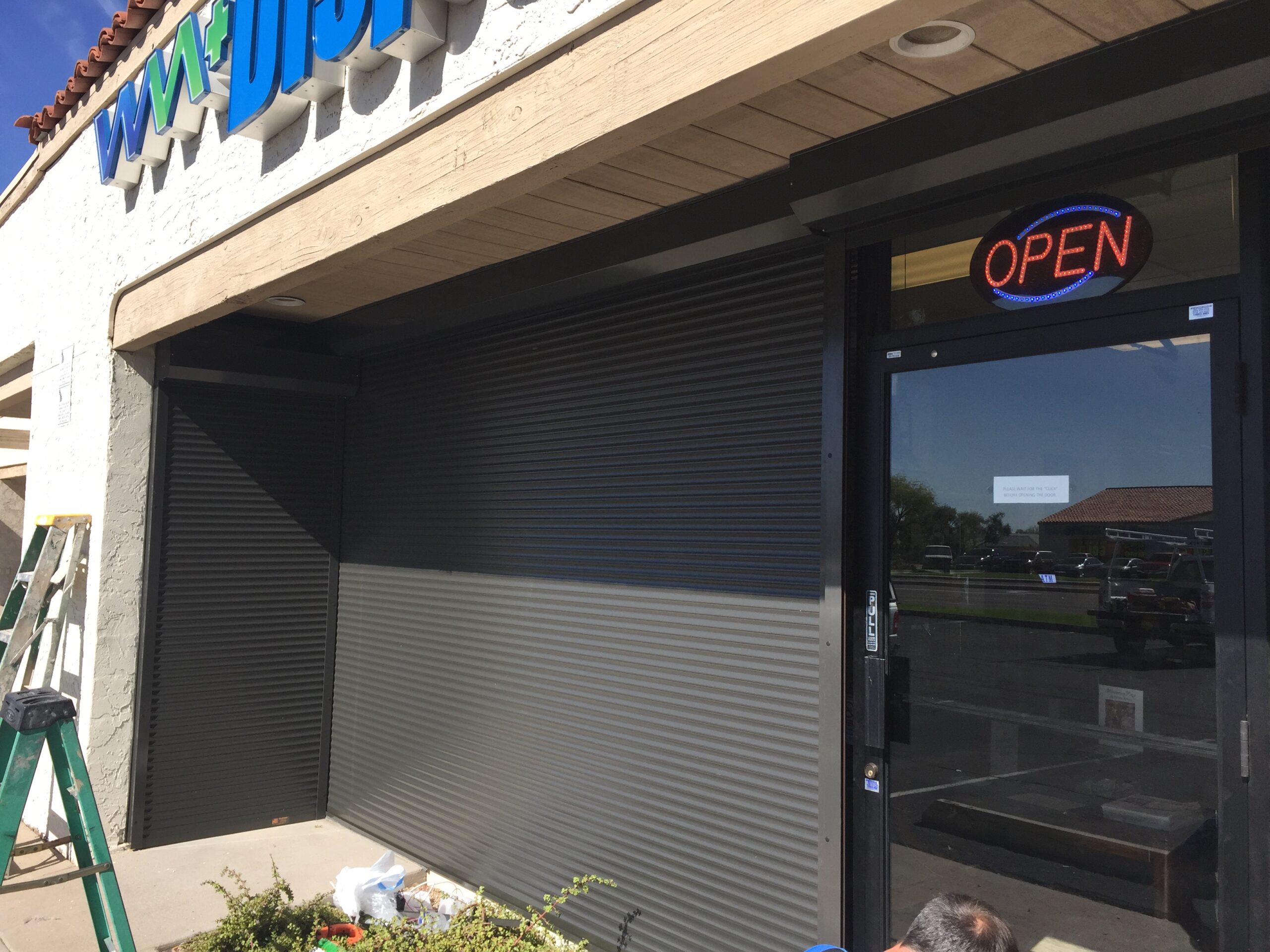 A storefront featuring a gray metal shutter partially closed, with a bright 'OPEN' sign displayed. The entrance area includes a small table with potted plants and part of a ladder visible on the left.