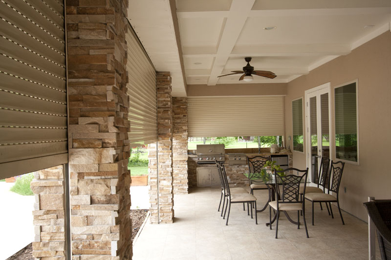 A covered outdoor patio area featuring stone columns, a dining set with metal chairs, a small table with flowers, and an outdoor kitchen visible in the background.