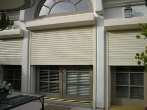 Exterior view of a house with three shuttered windows, featuring beige roll-down shutters and glass doors beneath. A small welcome mat is positioned in front of the doors.