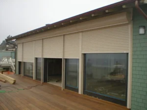 Exterior view of a modern house featuring large glass windows covered by beige roller shutters, with a wooden deck in the foreground and a gray sky above.