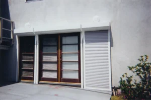 A pair of wooden double doors with glass panels, flanked by white rolling shutters, set against a light-colored wall. A small leafy plant is visible in the foreground on a concrete surface.