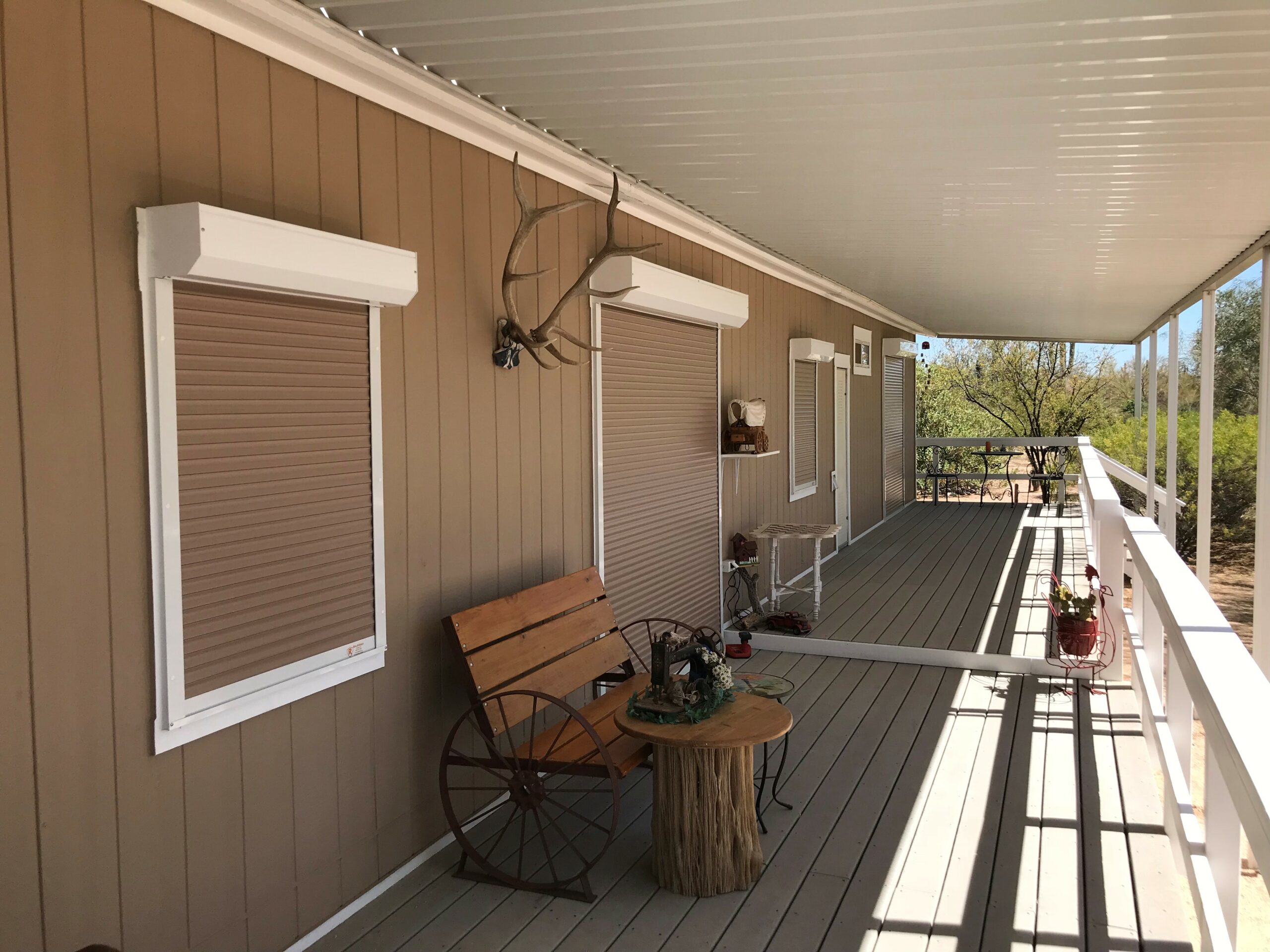 A wooden porch with two window shutters and a small wooden chair next to a round table. A decorative antler hangs above the windows, and a rustic wagon wheel is visible on the floor.