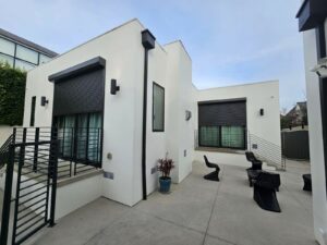 A contemporary white house with large windows and rolling shutters, featuring a modern staircase and outdoor seating area on a concrete patio.