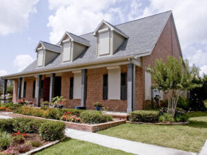 A two-story brick house with a pitched roof and multiple gabled dormers. The front yard features landscaped flower beds and a concrete pathway leading to the entrance.