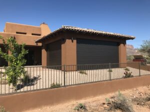 A modern brown house with a tiled roof and large black roller shutters, surrounded by a low fence and desert landscaping featuring sparse plants and gravel.