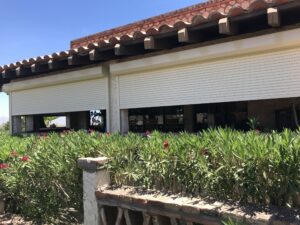 A house with a tiled roof features closed roller shutters on large windows, surrounded by green foliage and blooming pink flowers. The area is sunny and clear.