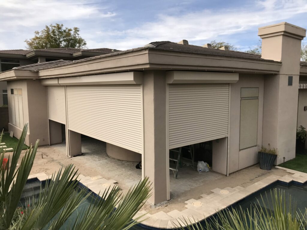 socal-pation-enclosures-2 A modern house exterior featuring beige walls and a flat roof. The garage is fitted with a large rolling shutter door. In the foreground, there is a landscaped area with plants and a swimming pool, partially visible.