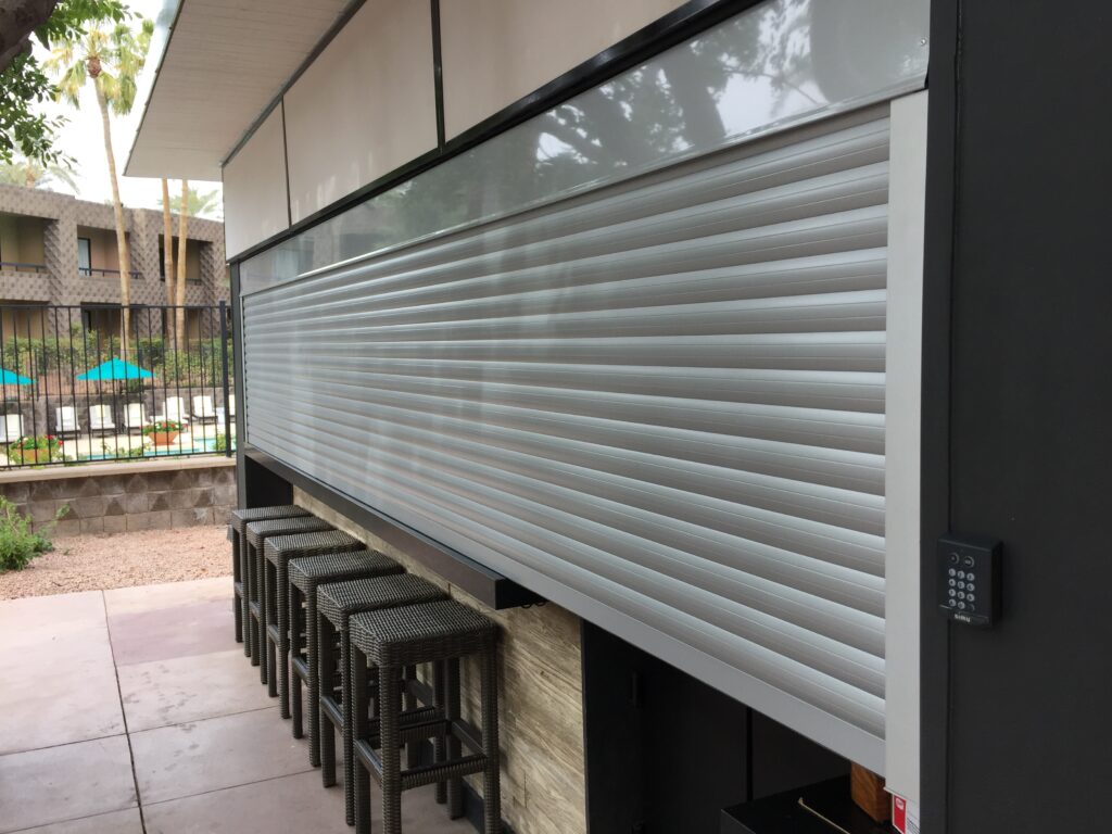 socal-shutter-store A modern bar area with a roll-up shutter closed. Four high stools are lined up in front of the bar. The background features a pool area with lounge chairs and trees.