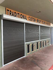 A closed tattoo shop with metal security shutters, featuring an orange sign above that reads 'TRADITIONAL • CUSTOM • BRIGHT COLORS • FINE DETAIL' and the word 'TATTOO' in bold letters.