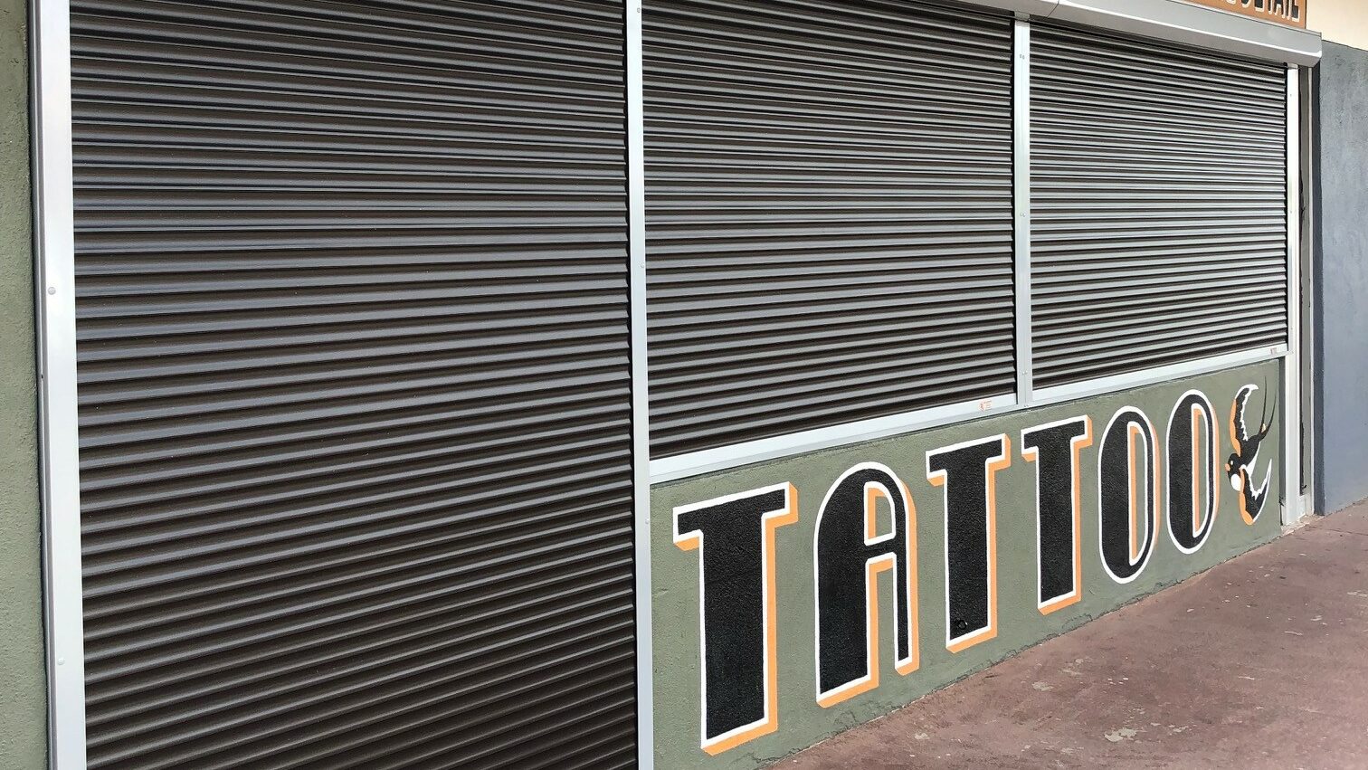 A closed tattoo shop with metal security shutters, featuring an orange sign above that reads 'TRADITIONAL • CUSTOM • BRIGHT COLORS • FINE DETAIL' and the word 'TATTOO' in bold letters.