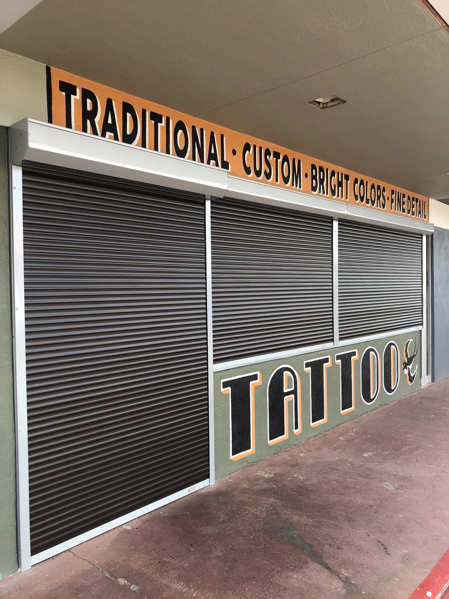 A closed tattoo shop with metal security shutters, featuring an orange sign above that reads 'TRADITIONAL • CUSTOM • BRIGHT COLORS • FINE DETAIL' and the word 'TATTOO' in bold letters.