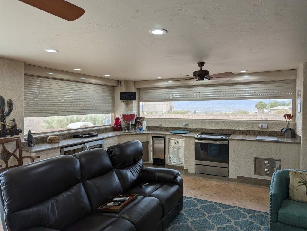 socal-sun-screen A modern kitchen and living area with a black leather couch in the foreground. The kitchen features a stove, refrigerator, and large windows with shades, providing a view of the outdoors.