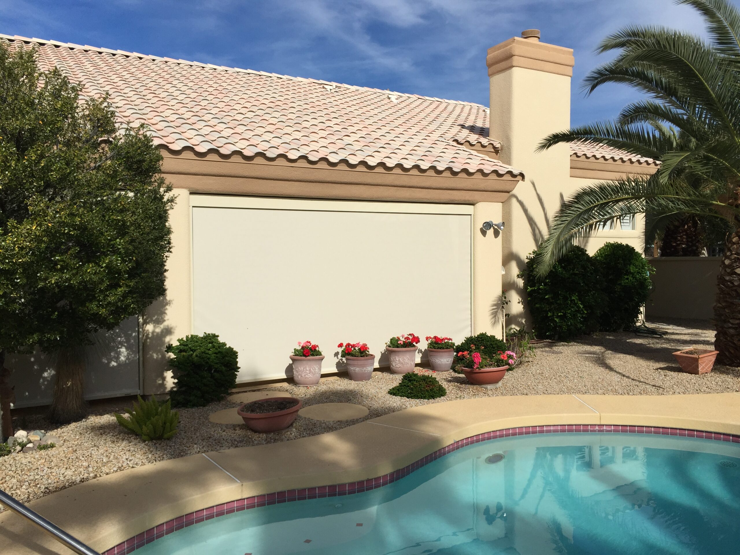 A residential house with a cream-colored exterior and a tiled roof. In front, there is a swimming pool and potted plants with flowers. Palm trees and low shrubs are visible around the house.