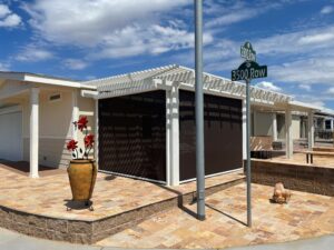 A residential building with a pergola and a dark screen enclosure, featuring a decorative pot with red flowers in front. A street sign indicates "3500 Row" nearby, under a blue sky with scattered clouds.