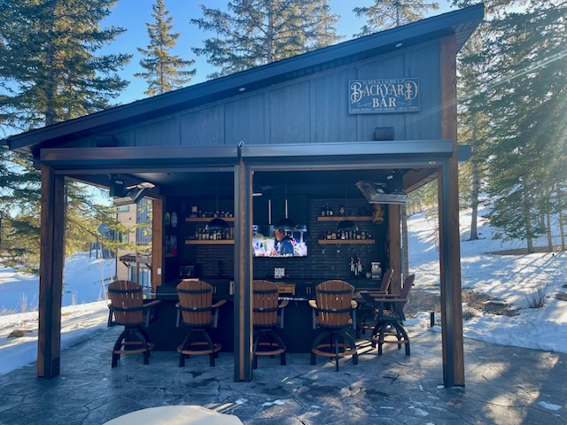A wooden backyard bar structure with a slanted roof, featuring bar stools in front. A television is mounted on the back wall, surrounded by shelves holding beverages. Snow covers the ground, and pine trees are visible in the background.