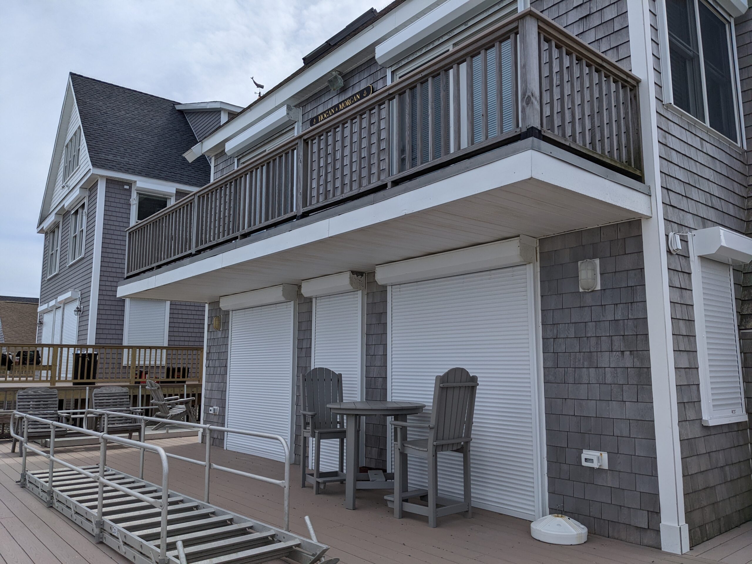 waterfront-3 A gray shingle-style beach house with a second-floor balcony. Below, there are three closed roll-up window shutters and a small outdoor table with chairs. Wooden decking is visible in the foreground.