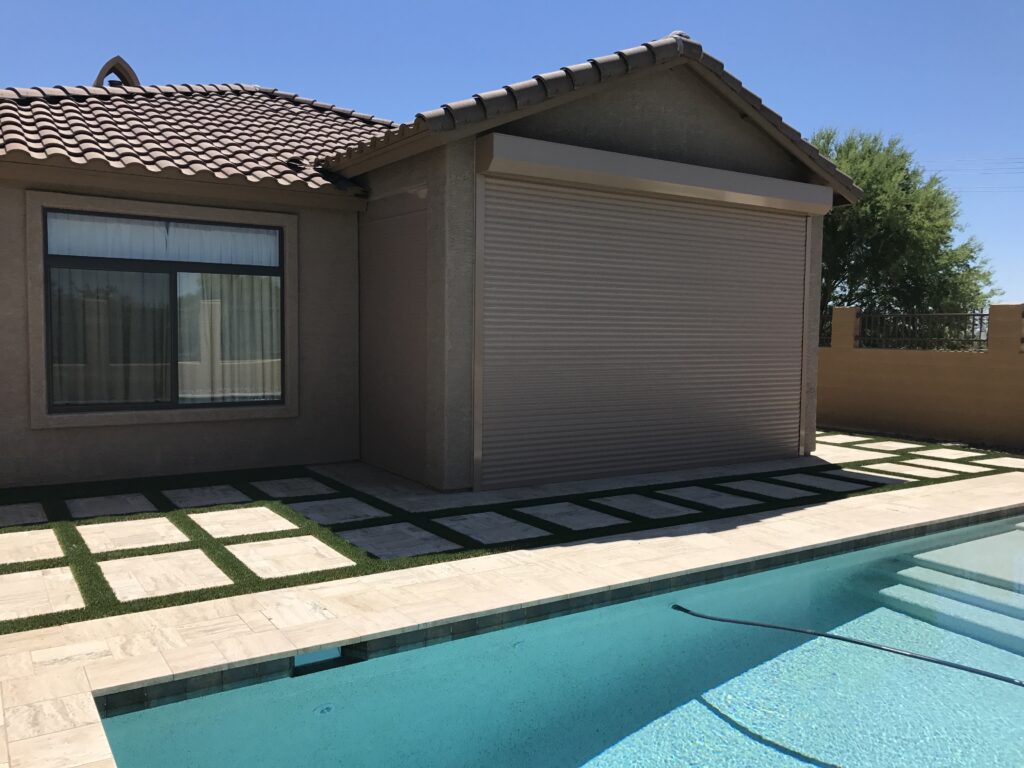 waterfront-5 A swimming pool adjacent to a beige house with a tiled roof. The house features a large window and a roll-up door. Green grass patches separate the stone pathway leading from the pool to the house.
