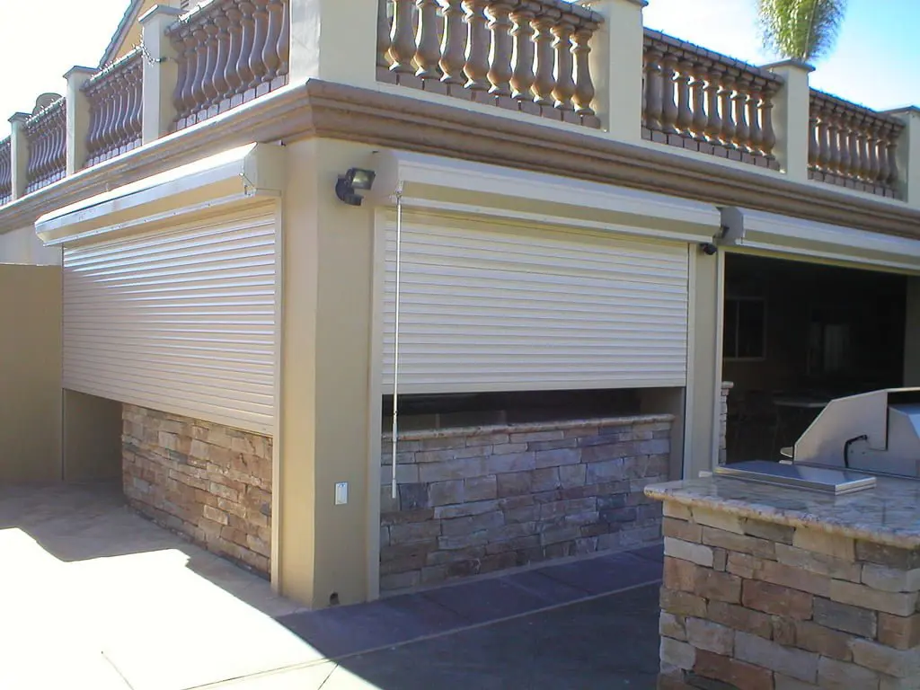 An exterior view of a house with a stone wall and beige roller shutters partially closed. The setting includes a patio area and a barbecue grill.