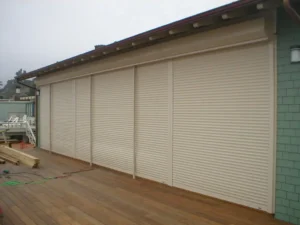 A wooden deck with several closed beige roll-up shutters mounted on a wall, partially obscuring a green building behind them, under a cloudy sky.