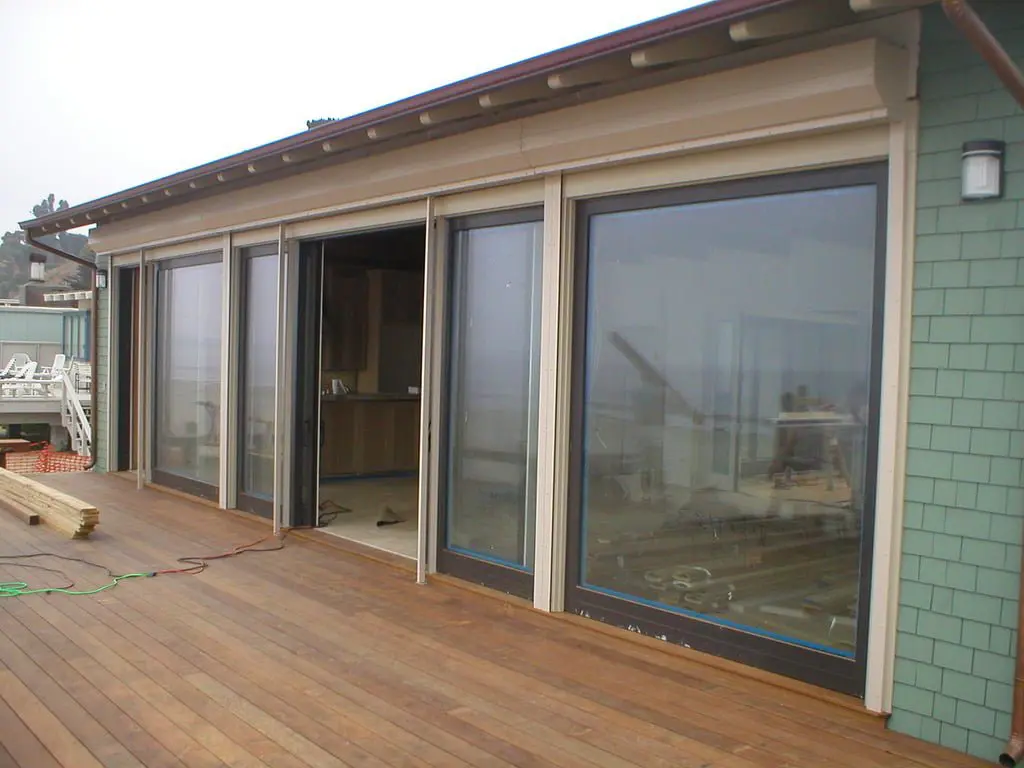 A modern house exterior featuring large sliding glass doors opening to a wooden deck. The structure has a green shingle wall and a flat roof, with construction materials visible nearby.
