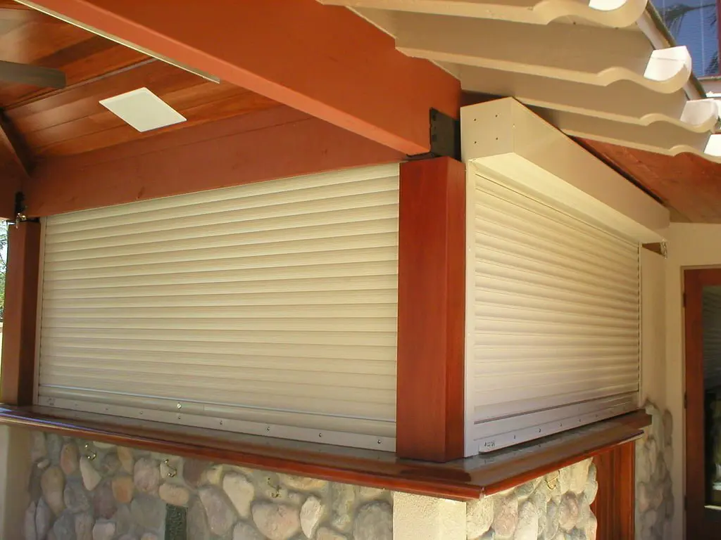 A partially closed rolling shutter mounted on the exterior of a building. The frame features wood accents, and the shutter is a light beige color set against a stone wall.