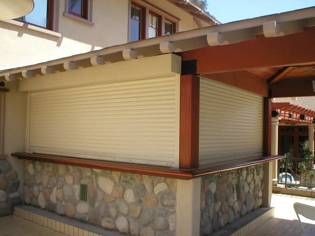 A closed roll-up shutter on a stone wall, part of a modern outdoor area with a wooden overhang. Nearby, there are windows and patio furniture visible in the background.
