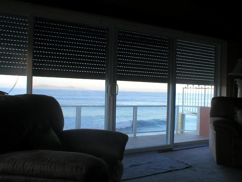 View of the ocean from inside a room, with large glass doors partially obscured by horizontal blinds, and a cushioned chair visible in the foreground.