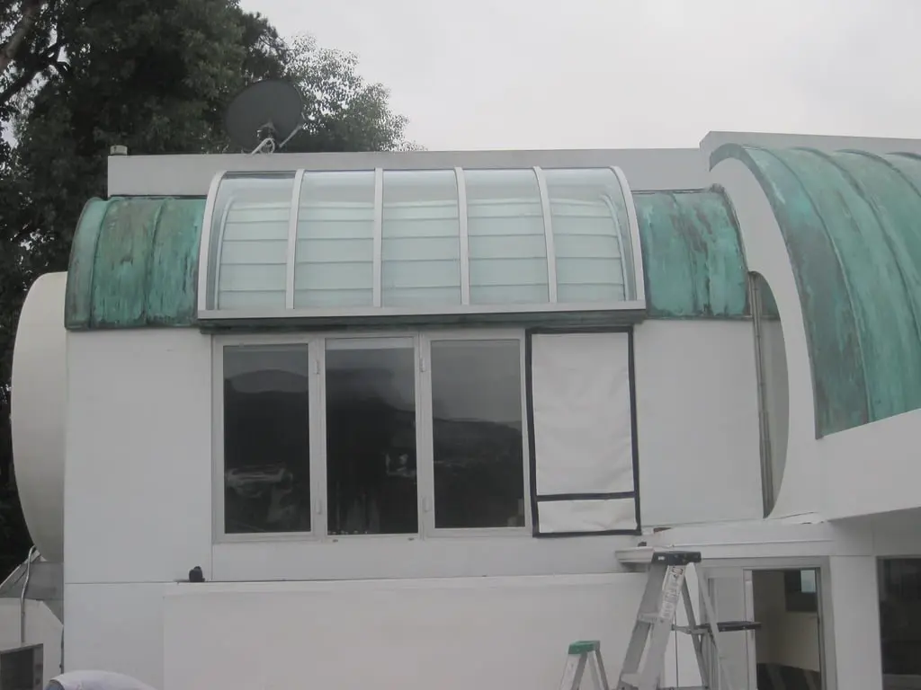 A modern building with a curved roof featuring a glass skylight and green copper accents. A small ladder leans against the side. The sky is overcast, indicating a cloudy day.