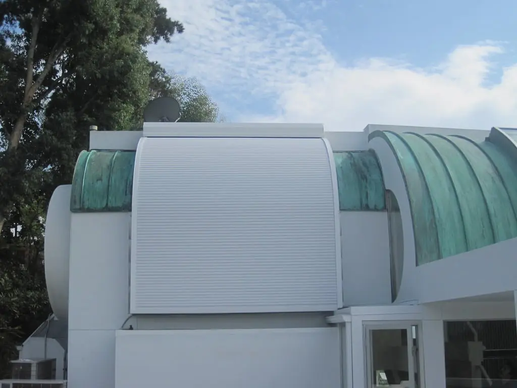 A modern building exterior with an arched green roof and a large white roller shutter partially closed. The structure is surrounded by trees under a partly cloudy sky.