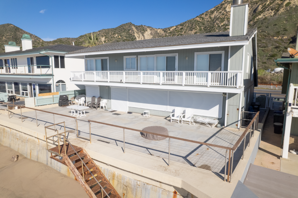 Exterior_Drone_01 A modern beachfront house with a large deck and patio furniture, situated next to a narrow sand area. The house features a gray exterior and a balcony overlooking the beach, with mountains visible in the background.